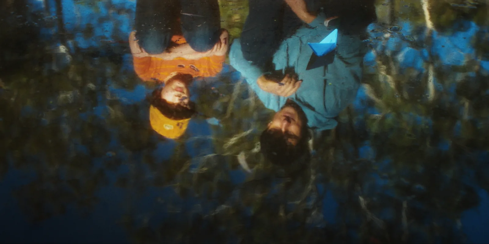 Two boys reflected upside down in a still pool, one in an orange shirt and yellow cap, the other holding a blue paper boat, gum trees mirrored around them.