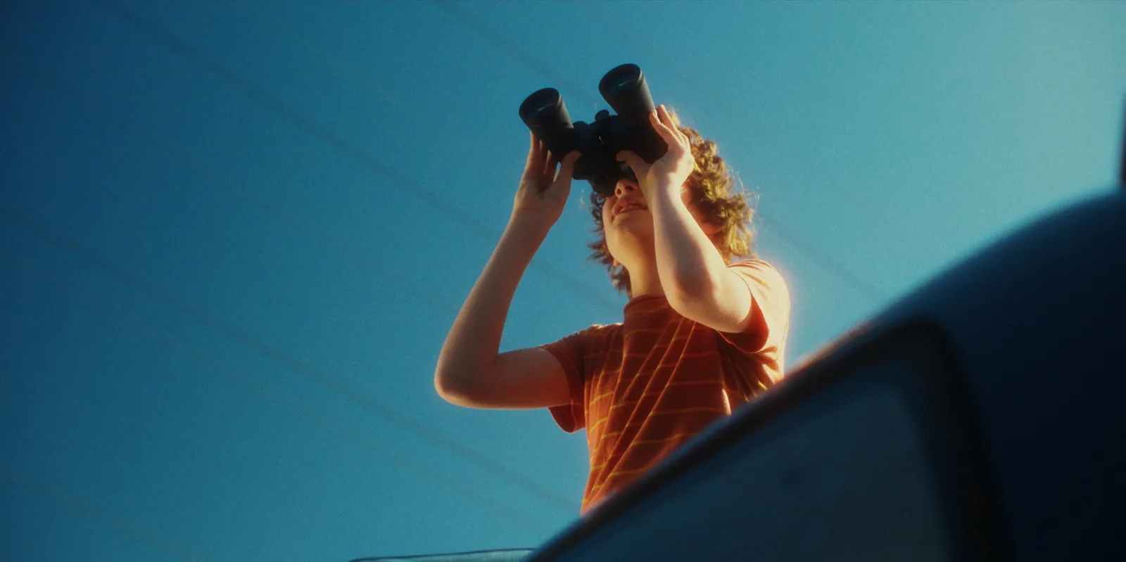 A curly-haired boy in a striped orange T-shirt looks up through binoculars against a clear blue sky, shot from below past the edge of a car.