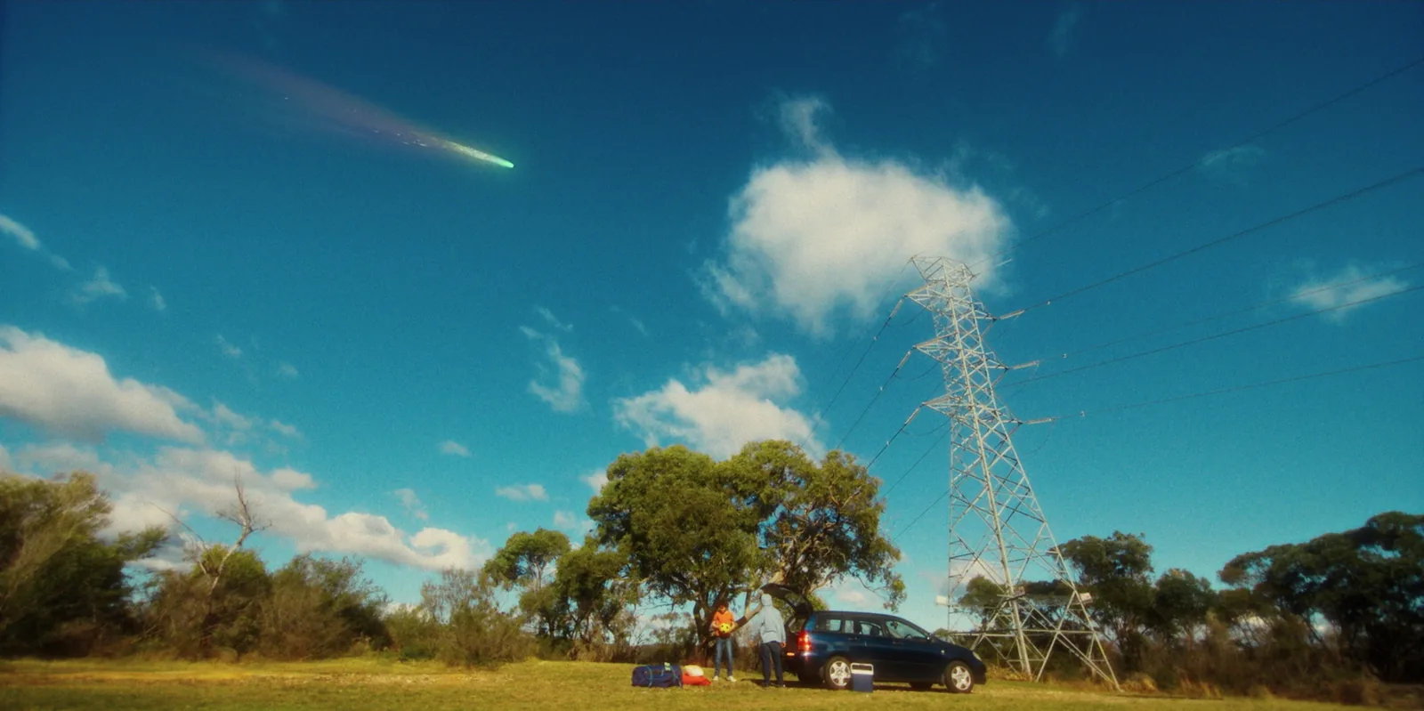 A wide field under a blue sky: a car with its boot open beside a power pylon, two figures by the car and a green-tailed comet streaking overhead.