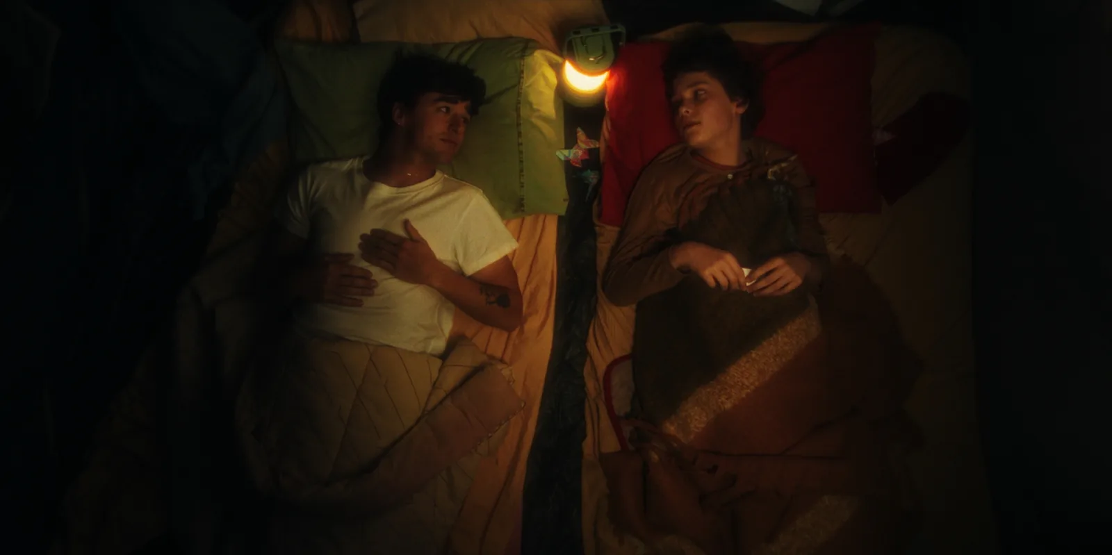Top-down on two young men lying in separate beds, one in a white T-shirt, lit by a small glowing lamp between them.