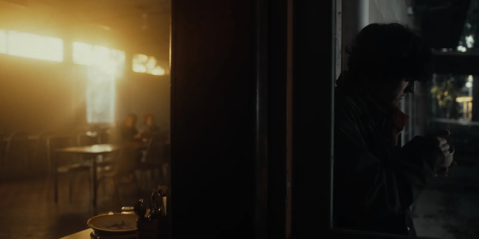 A man in a jacket stands in a dark doorway on the right while low sun pours through the windows of an empty diner on the left.