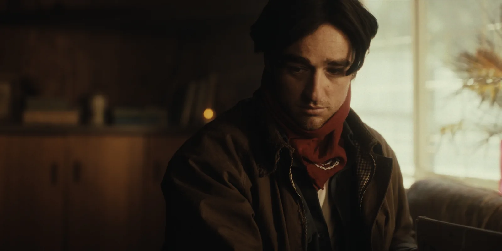 A young man with dark hair and a red bandana looks down, a bright window beside him and dim shelves behind.