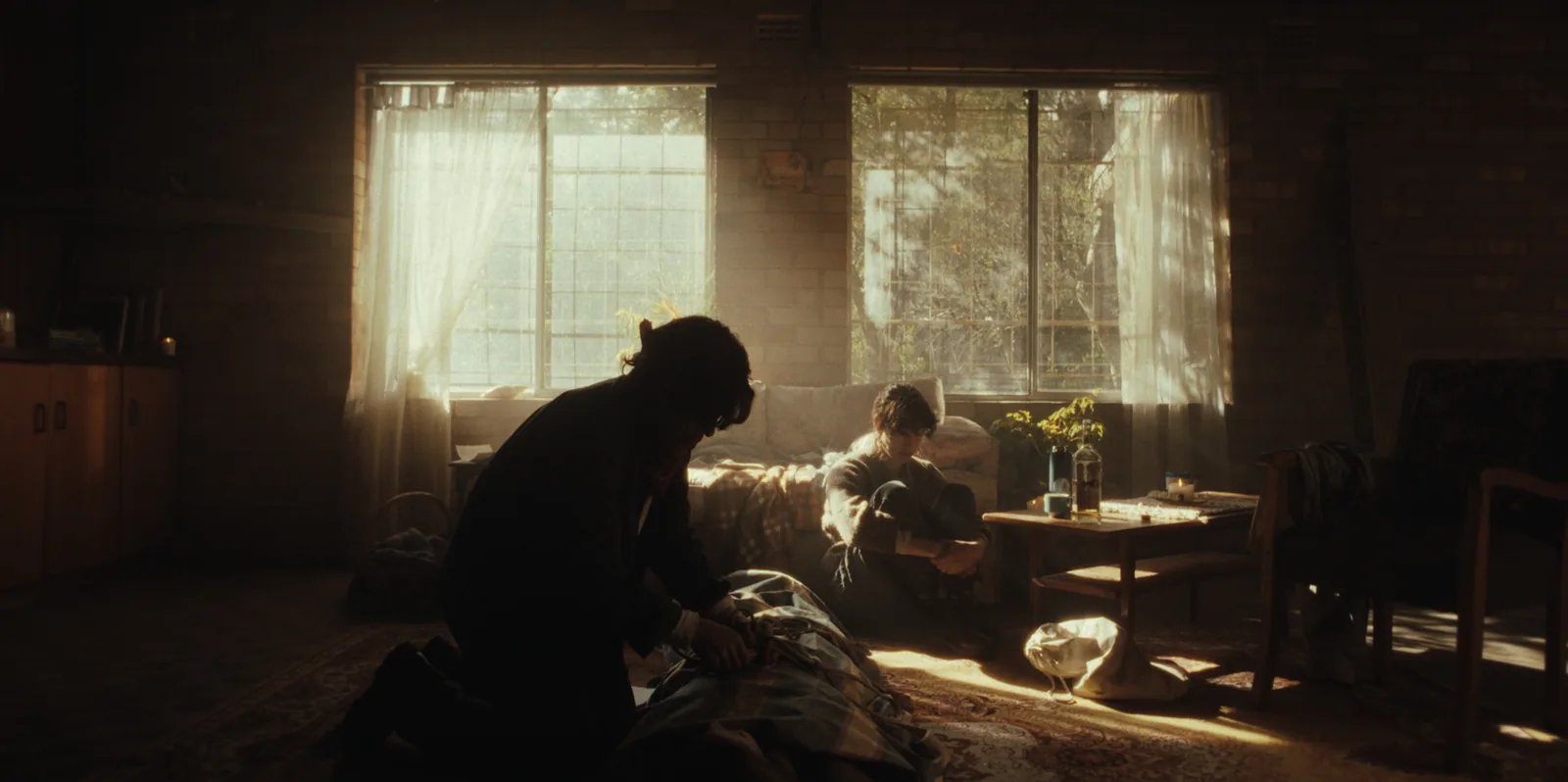 A sunlit living room seen against its windows: one figure kneels over a blanket on the floor while another sits by the sofa.