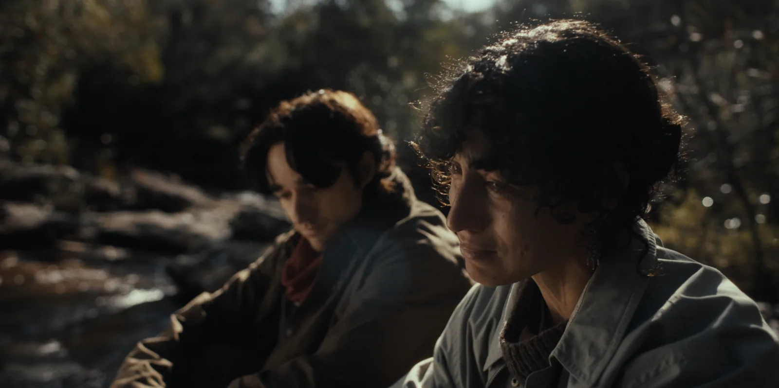 Two people sit by a rocky creek, a curly-haired woman sharp in the foreground and a young man soft behind her, backlit by the sun.
