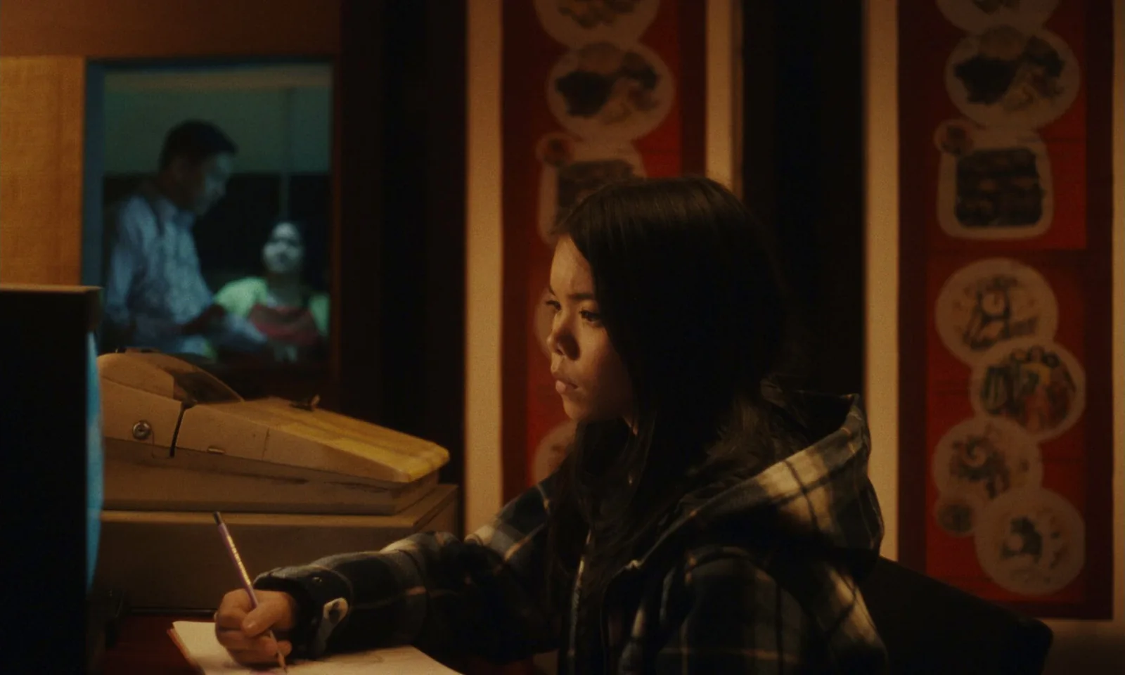 A girl in a hooded flannel shirt writes at a desk beside an old fax machine, a television behind her showing a man and a woman.