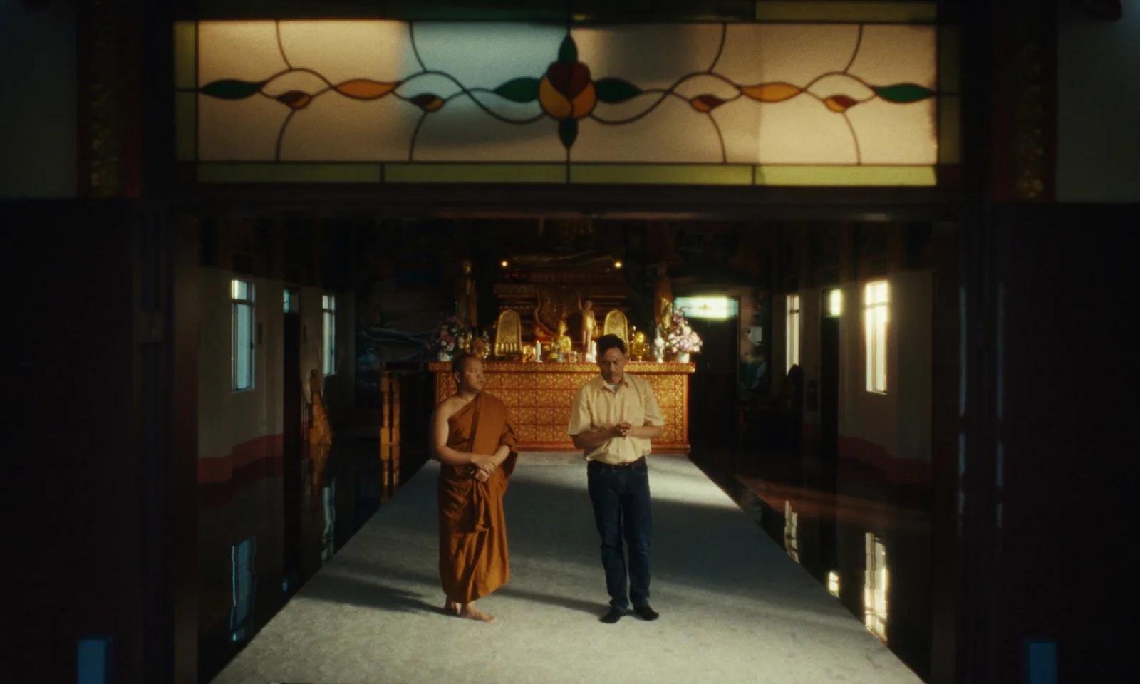 A monk in saffron robes and a man in a yellow shirt stand in a long temple hall under a leadlight window, a gold altar at the far end.