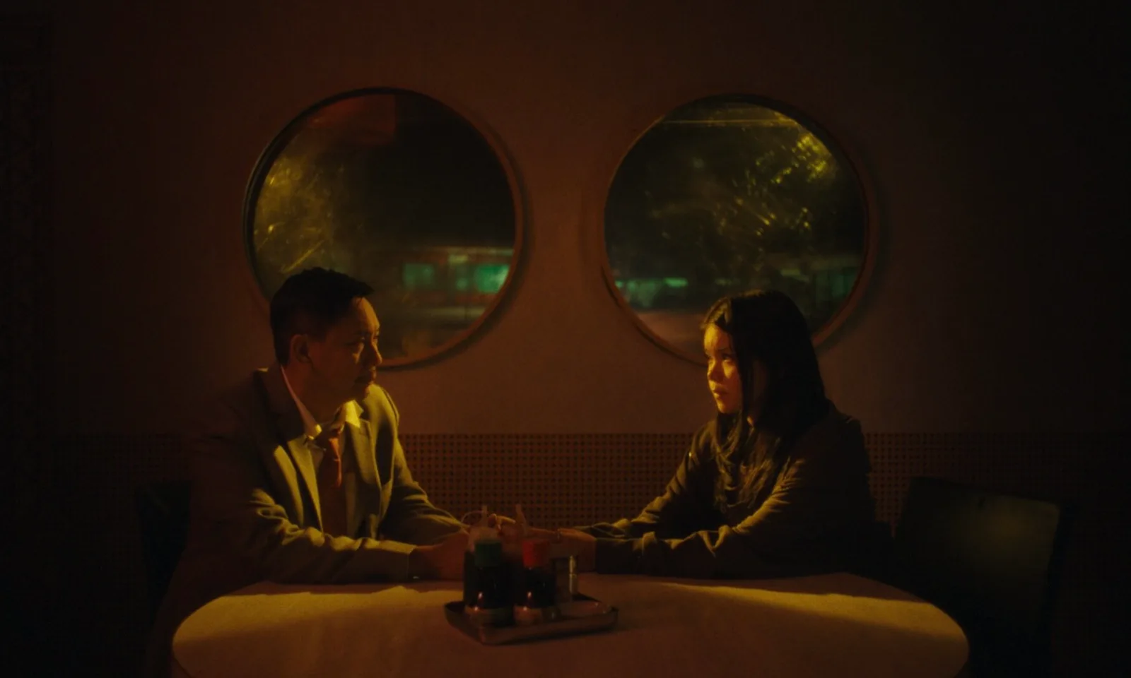 A man and a girl sit across a small table in a dim restaurant, two round windows glowing behind them.