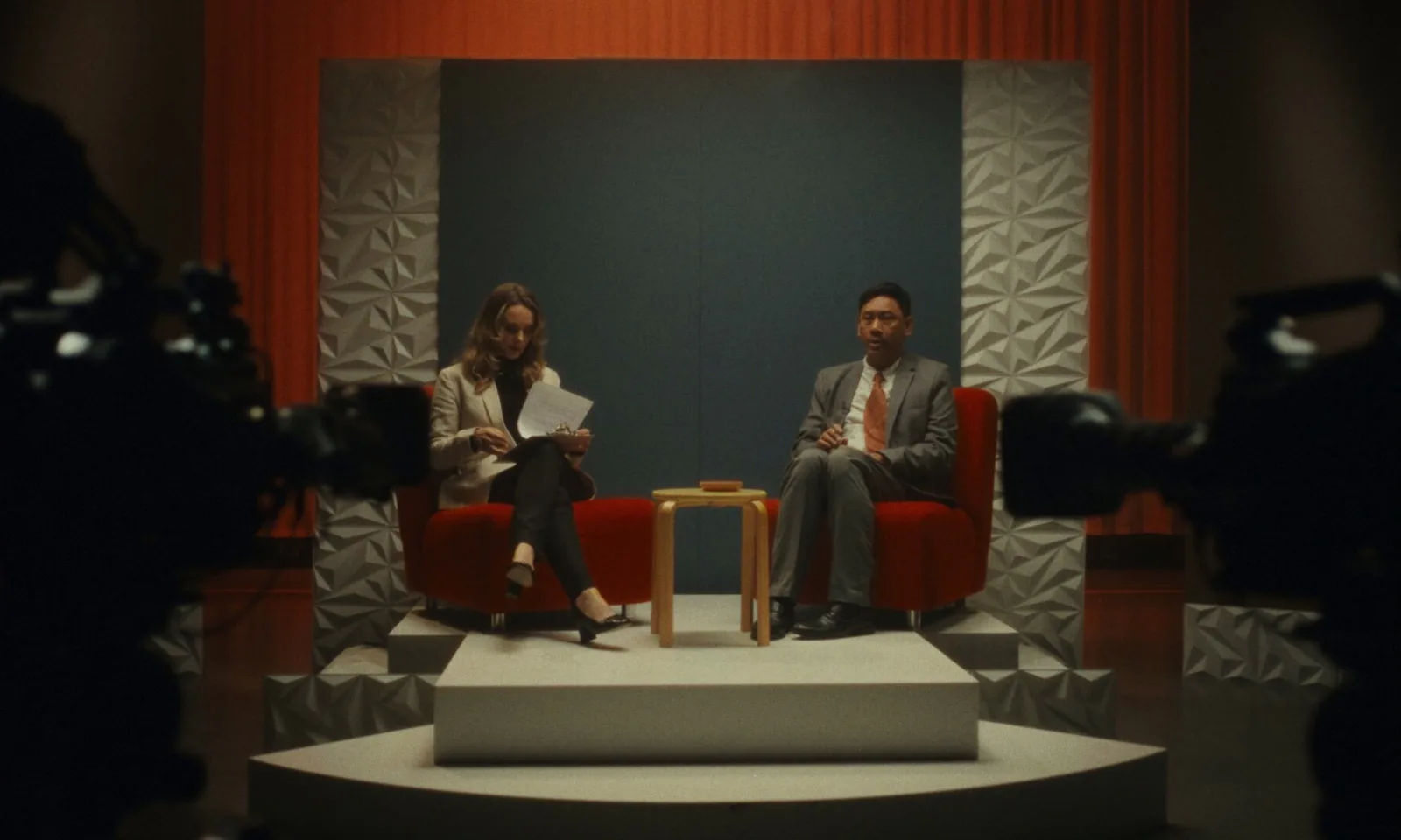 A television interview set: a woman with notes and a man in a grey suit on red armchairs, framed between two studio cameras in the foreground.