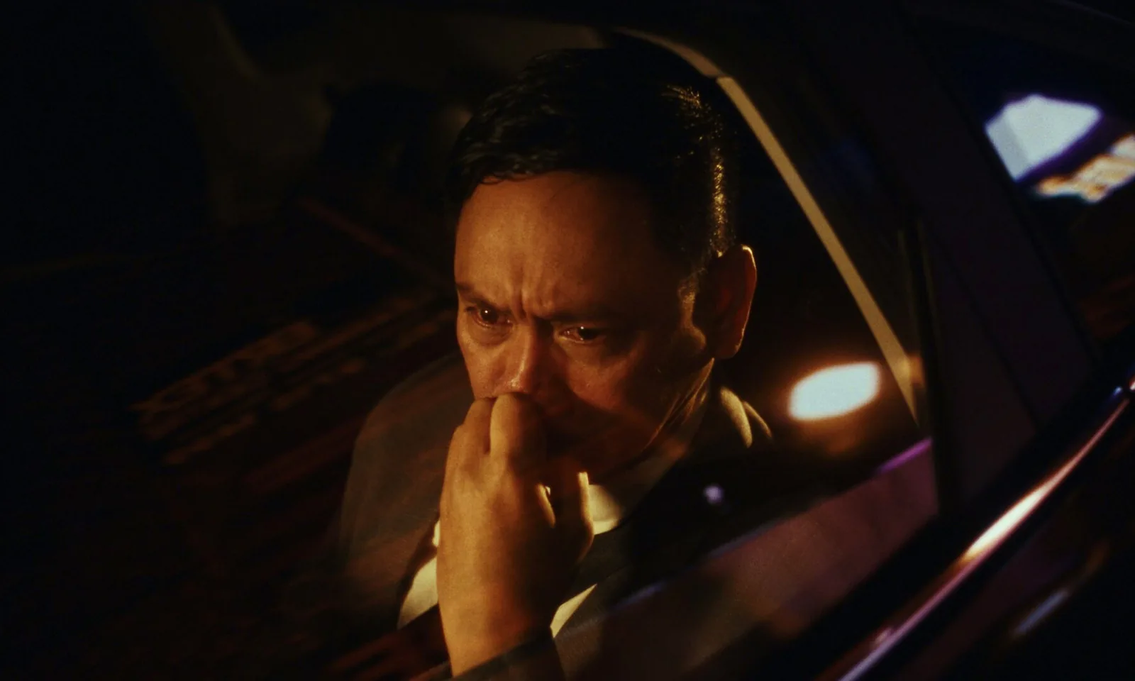 A man in a suit sits in a car at night with his fist to his mouth, lights reflected in the window beside him.