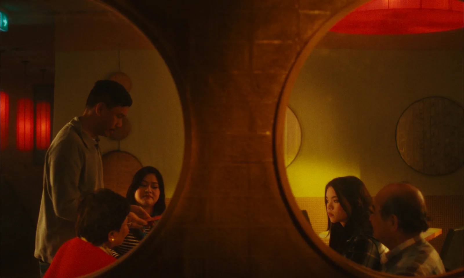 Two round openings in a wall look into a restaurant: a man leans over a family at one table, and a girl and an older man sit at another.