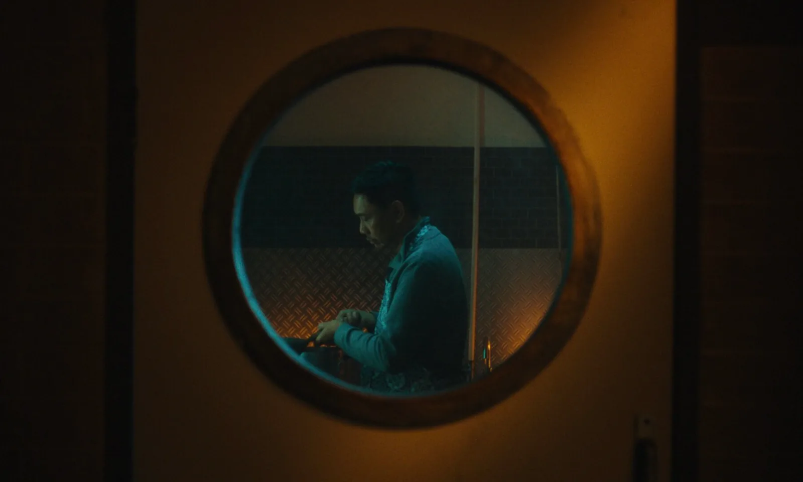 A man sits with his head down, seen through a round window set in a warm, dark wall.