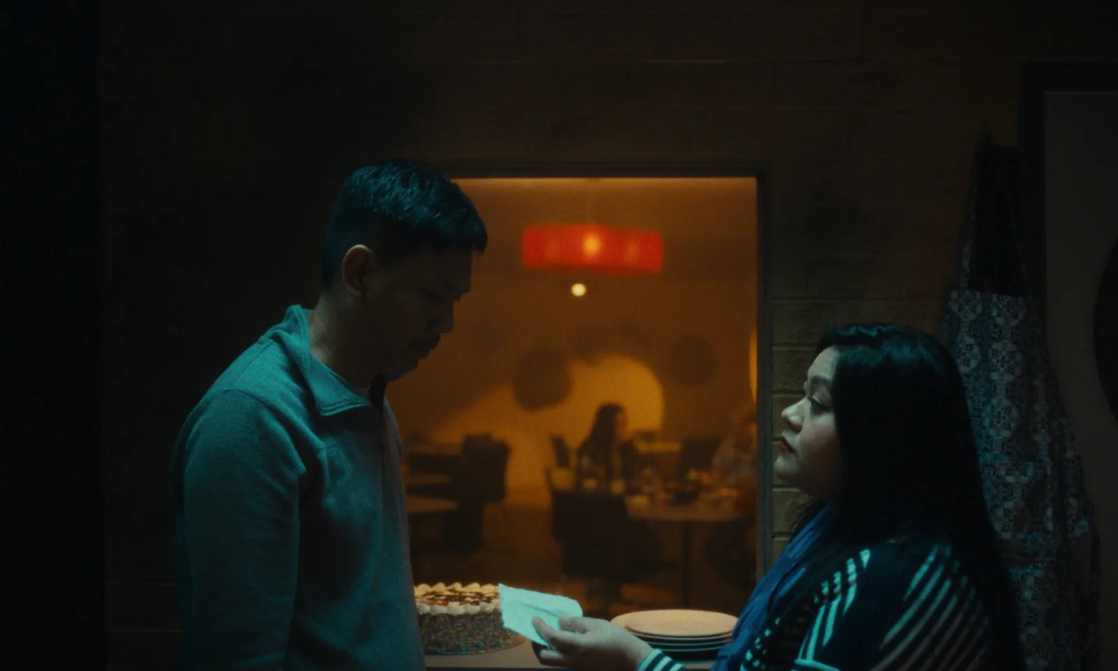 A man looks down as a woman hands him a note beside a cake, a restaurant glowing through the hatch behind them.