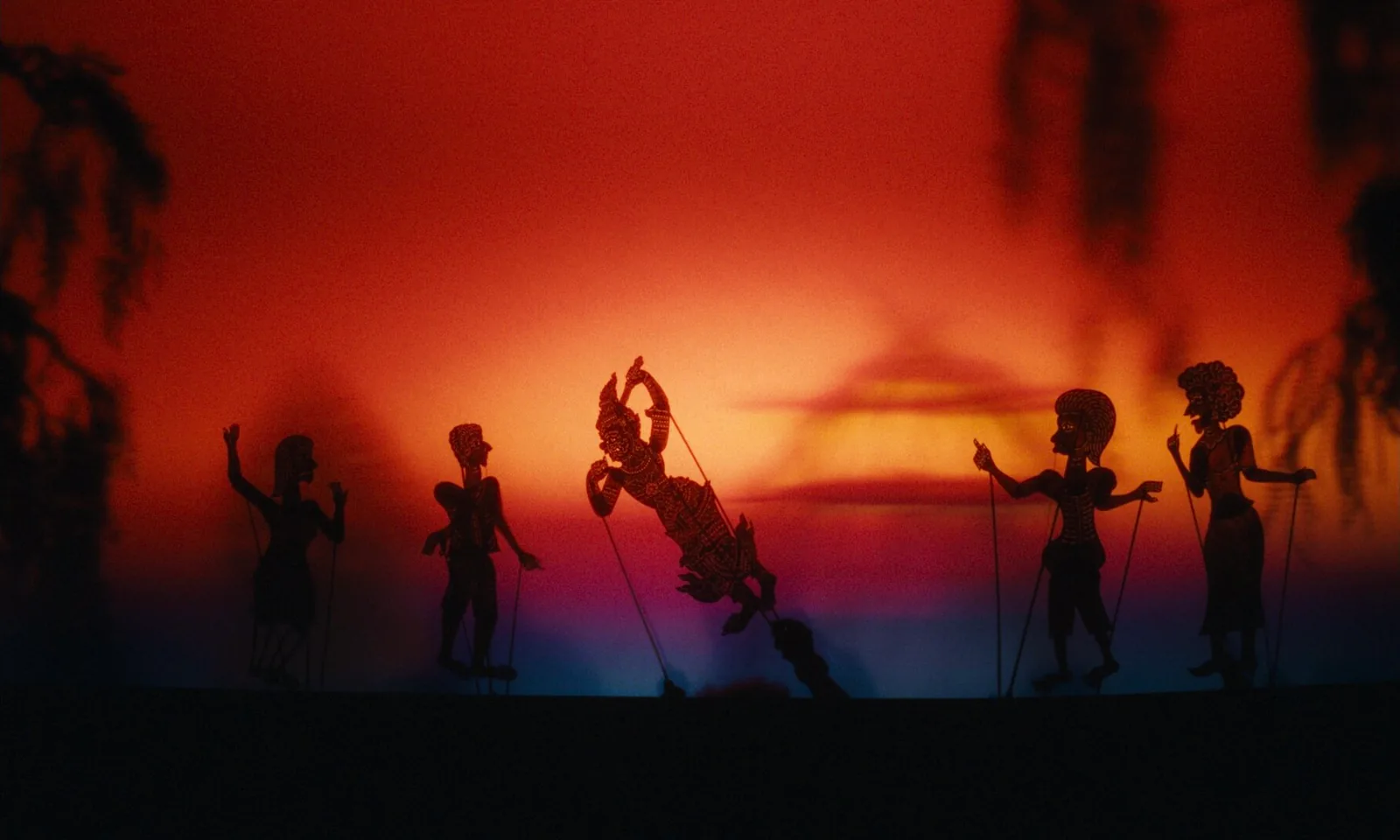 Shadow puppets silhouetted against a red and orange screen, soft leaves hanging in the foreground.