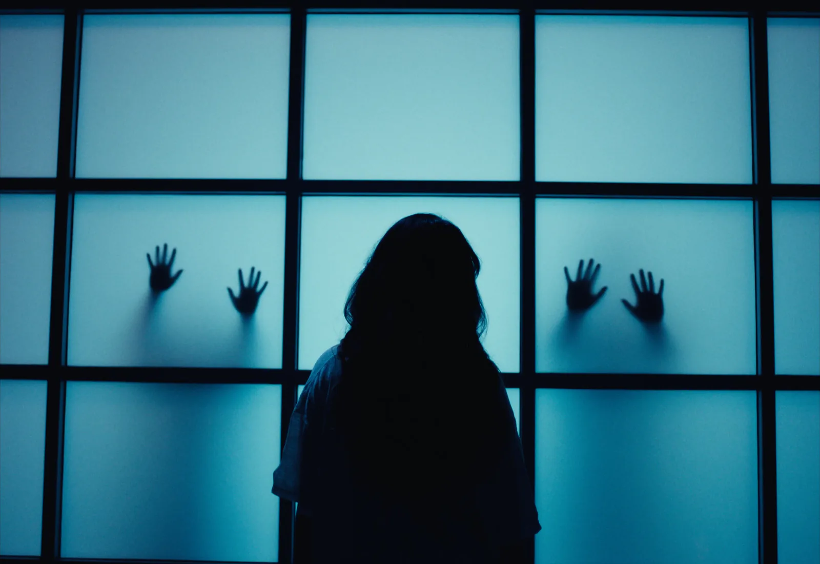 A long-haired figure in silhouette faces a backlit blue window while hands press against the glass.