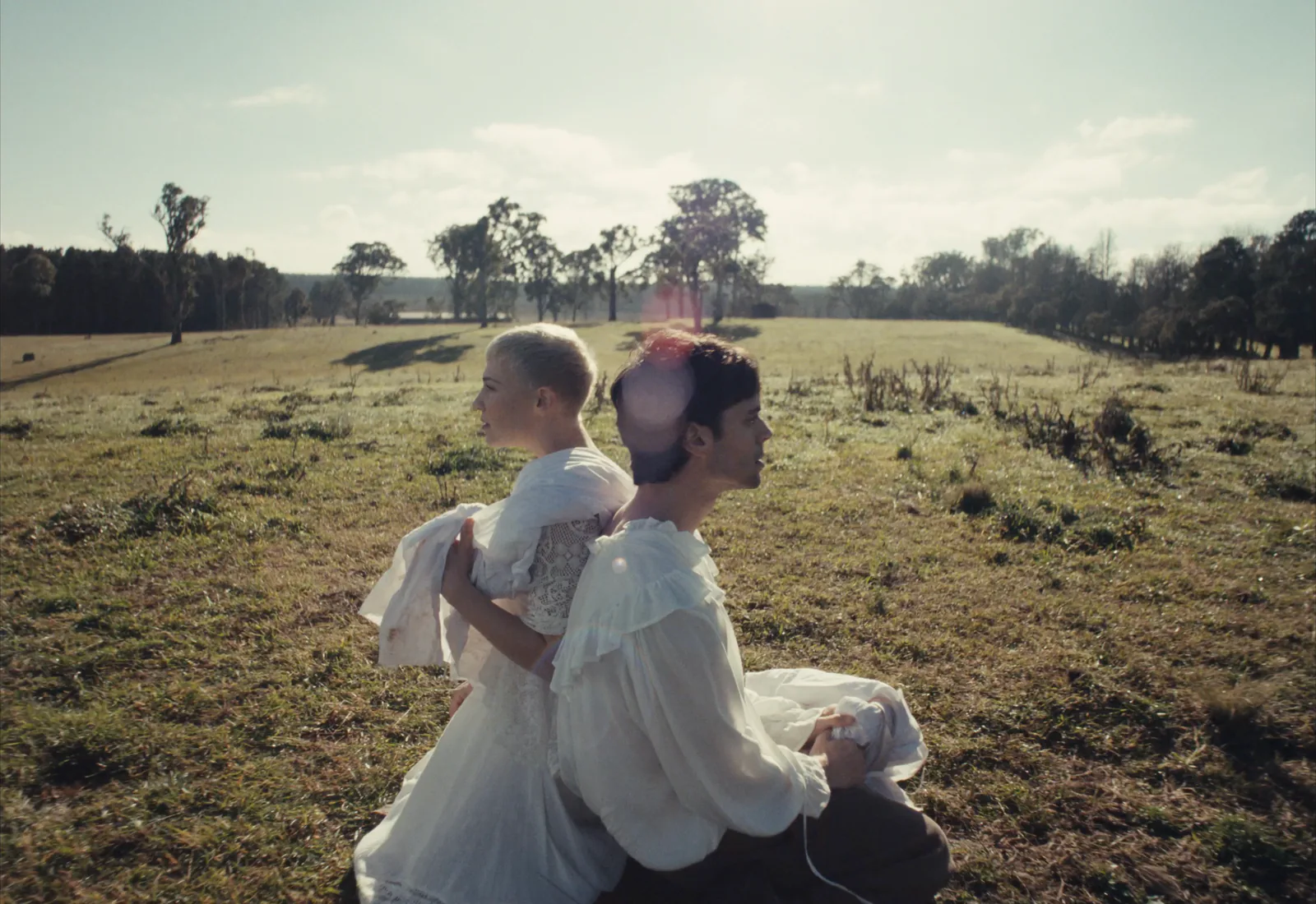 Two people in white period clothes sit back to back in a sunny paddock, a soft pink flare ghost over one head.