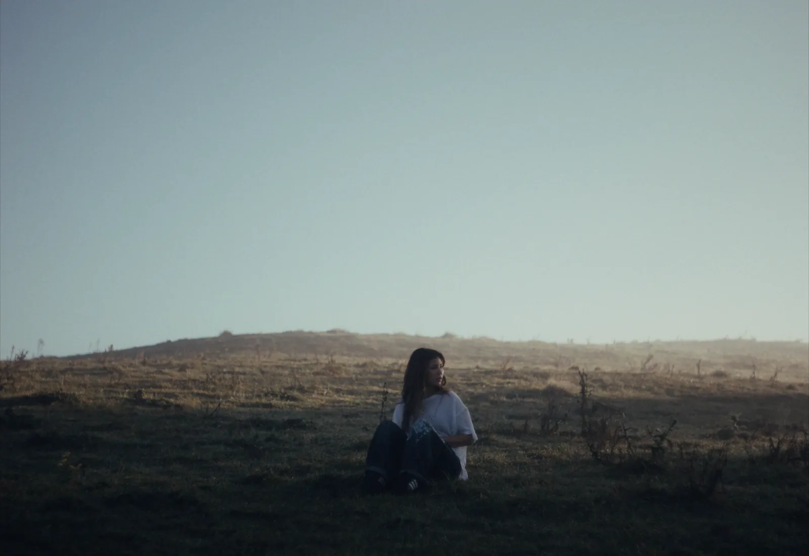 A woman sits alone in the grass below a misty hill, the backlit sky soft and milky.