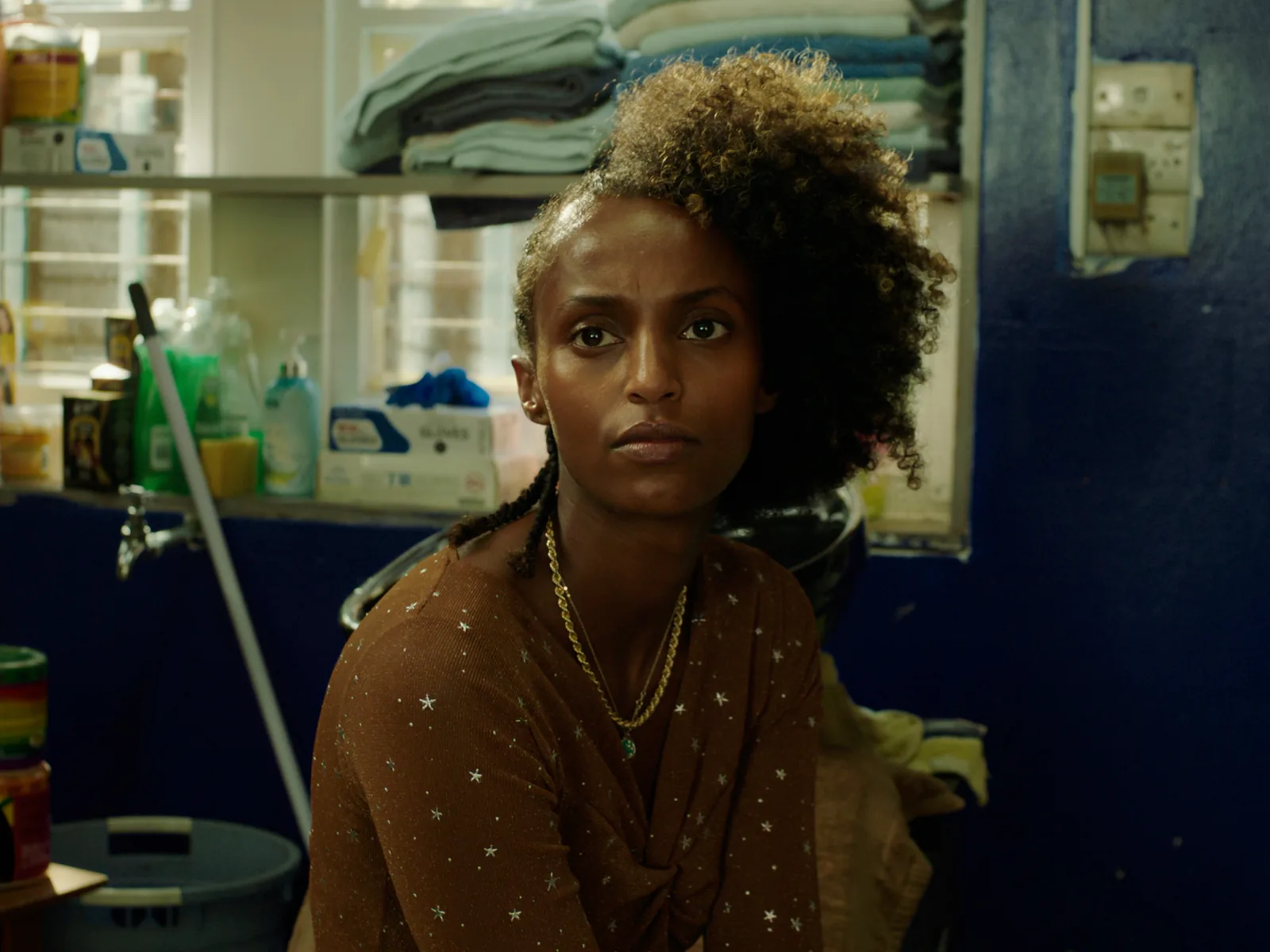 A young woman with curly hair and a star-print top sits in a salon and glances to one side, shelves of towels soft behind her.