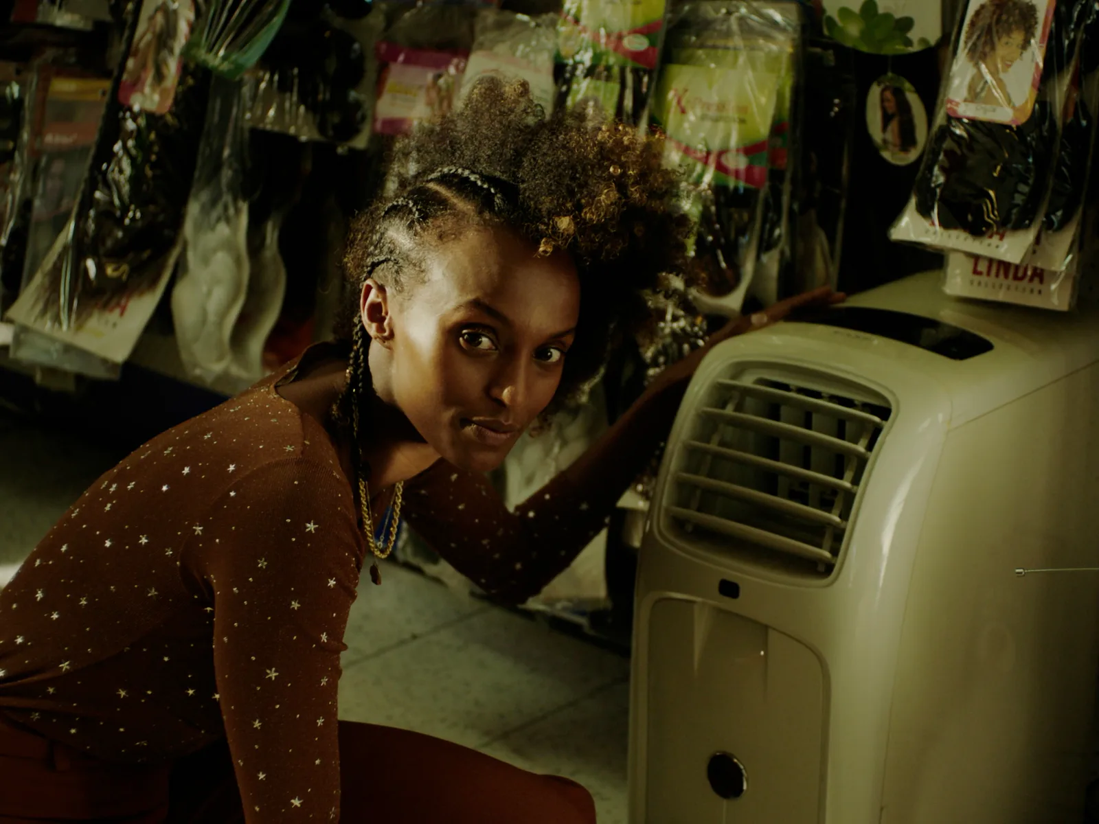 A young woman crouches beside a portable air conditioner in a salon, packets of hair extensions hanging on the wall behind her.