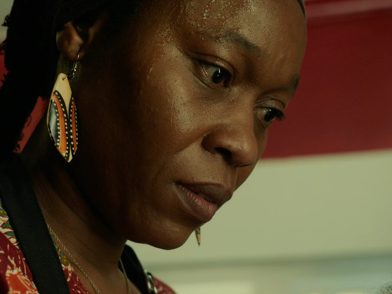 A tight close-up of an older woman with a painted wooden earring looking down, sweat on her face.