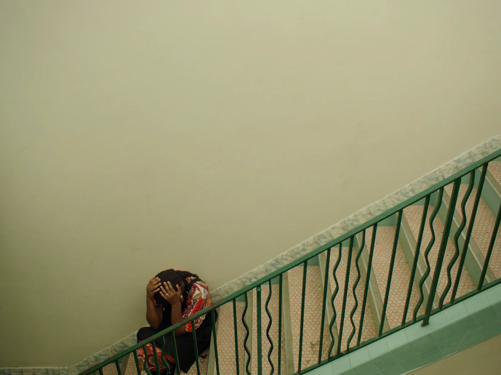 Looking down a stairwell with a green wavy railing, a figure sits hunched on the steps with both hands over their head.
