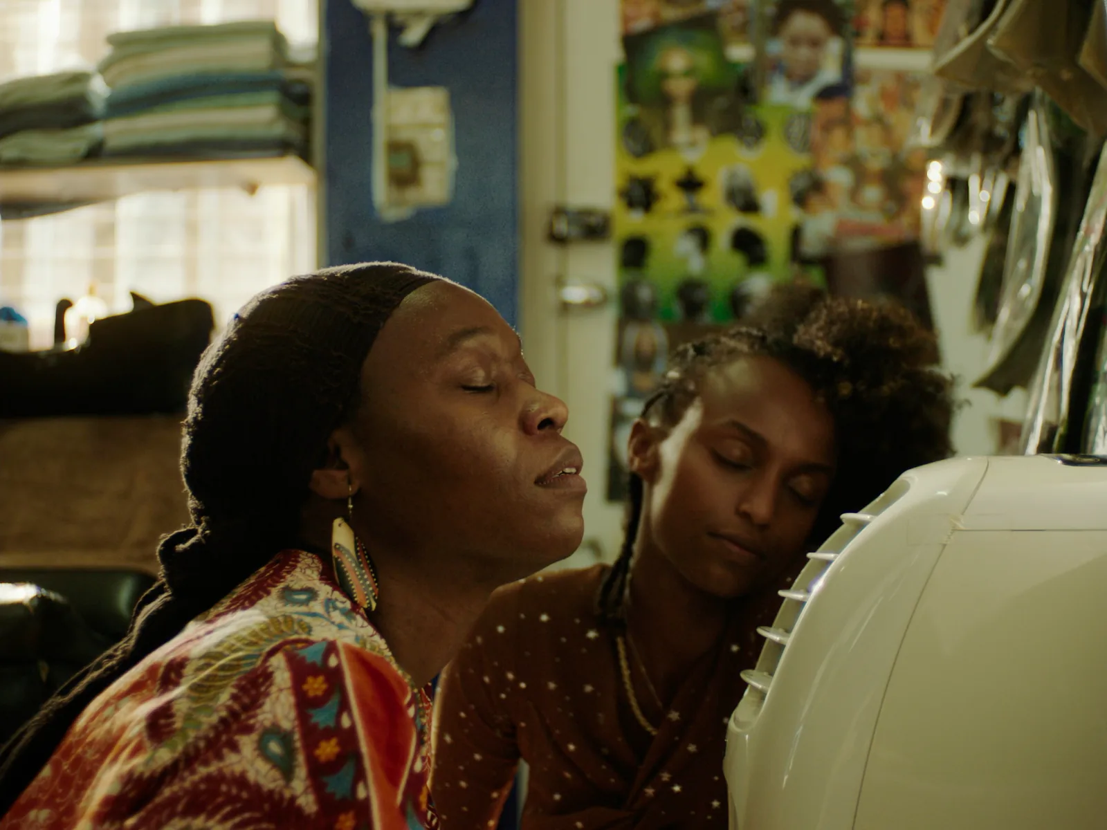 Two women lean in toward a portable air conditioner with their eyes closed, the older one sharp in front and the younger one soft behind.