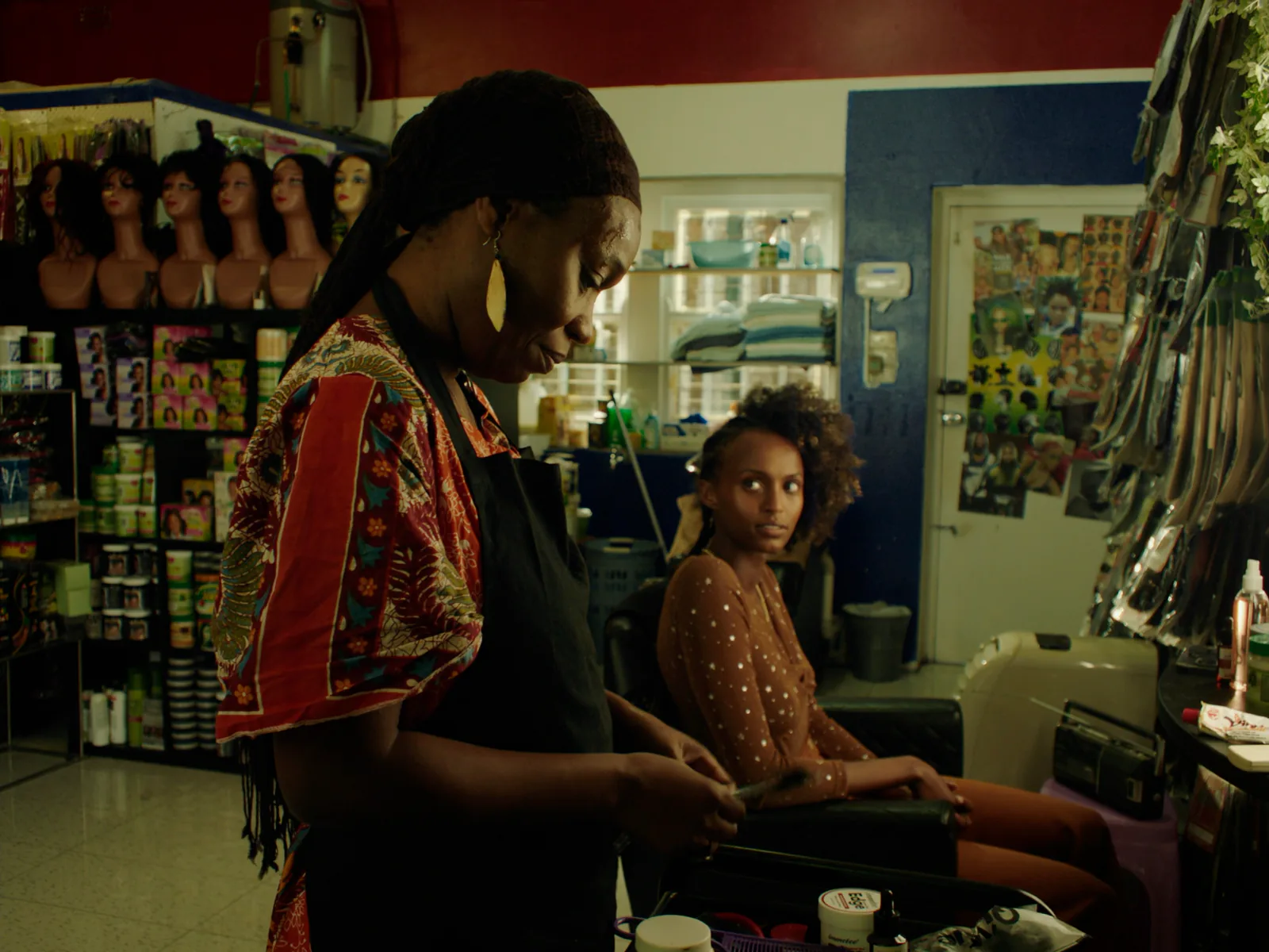 In a busy salon, a woman in an apron stands in the foreground while a younger woman sits waiting in a chair behind her.