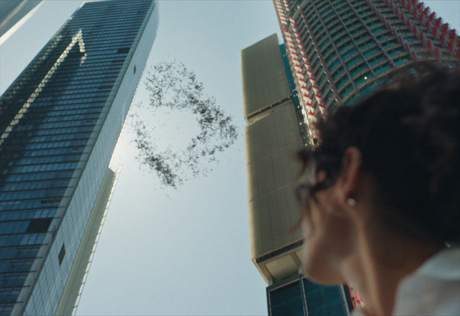 Looking straight up between two city towers at a flock of birds wheeling in a ring, a woman's curly hair soft in the foreground.