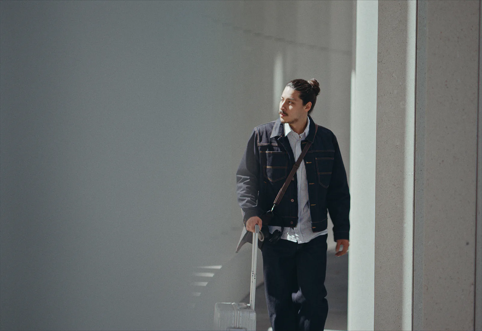 A man in a denim jacket wheels a suitcase down a bright white corridor, the walls soft around him.