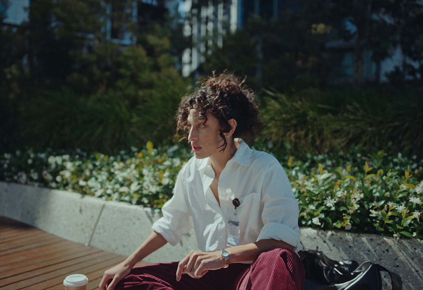 A woman with curly hair and a white shirt sits on a bench in a city garden in hard afternoon sun, plants behind her.