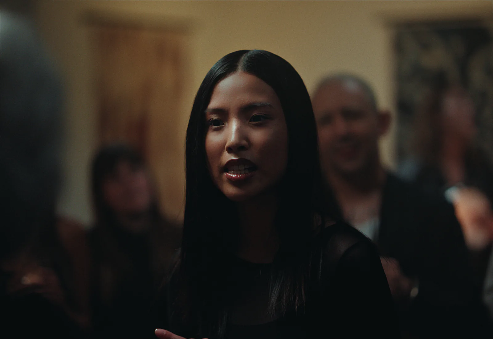 A woman with long dark hair talks at an evening event while the guests behind her fall soft into the warm room.