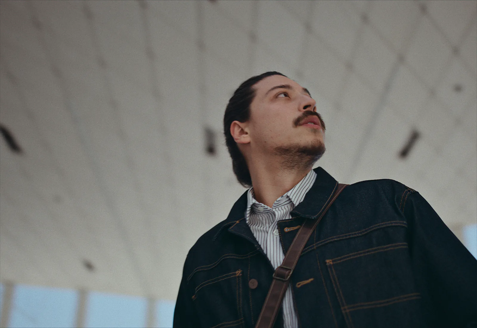 A man with a moustache and a denim jacket looks up and off to one side beneath a white panelled ceiling.