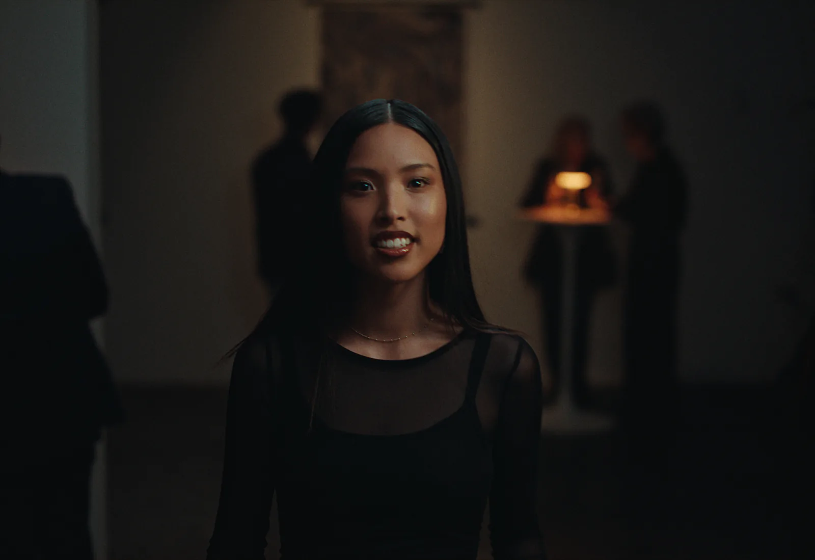 A smiling woman in a sheer black top stands in a dim room, a small lamp glowing on a table among the guests behind her.